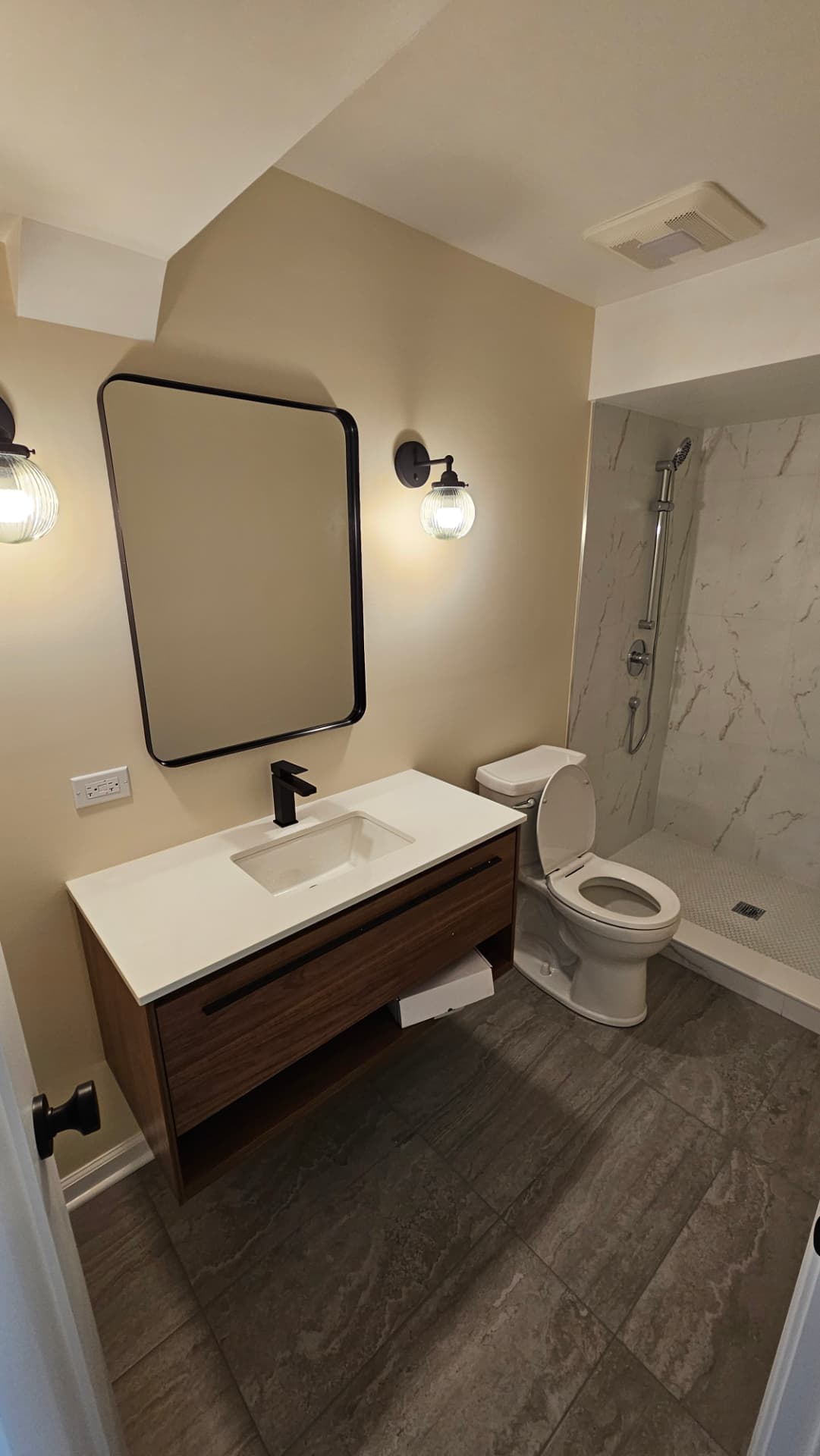 Bathroom with a floating walnut vanity, black framed mirror, glass sconces and a tiled shower